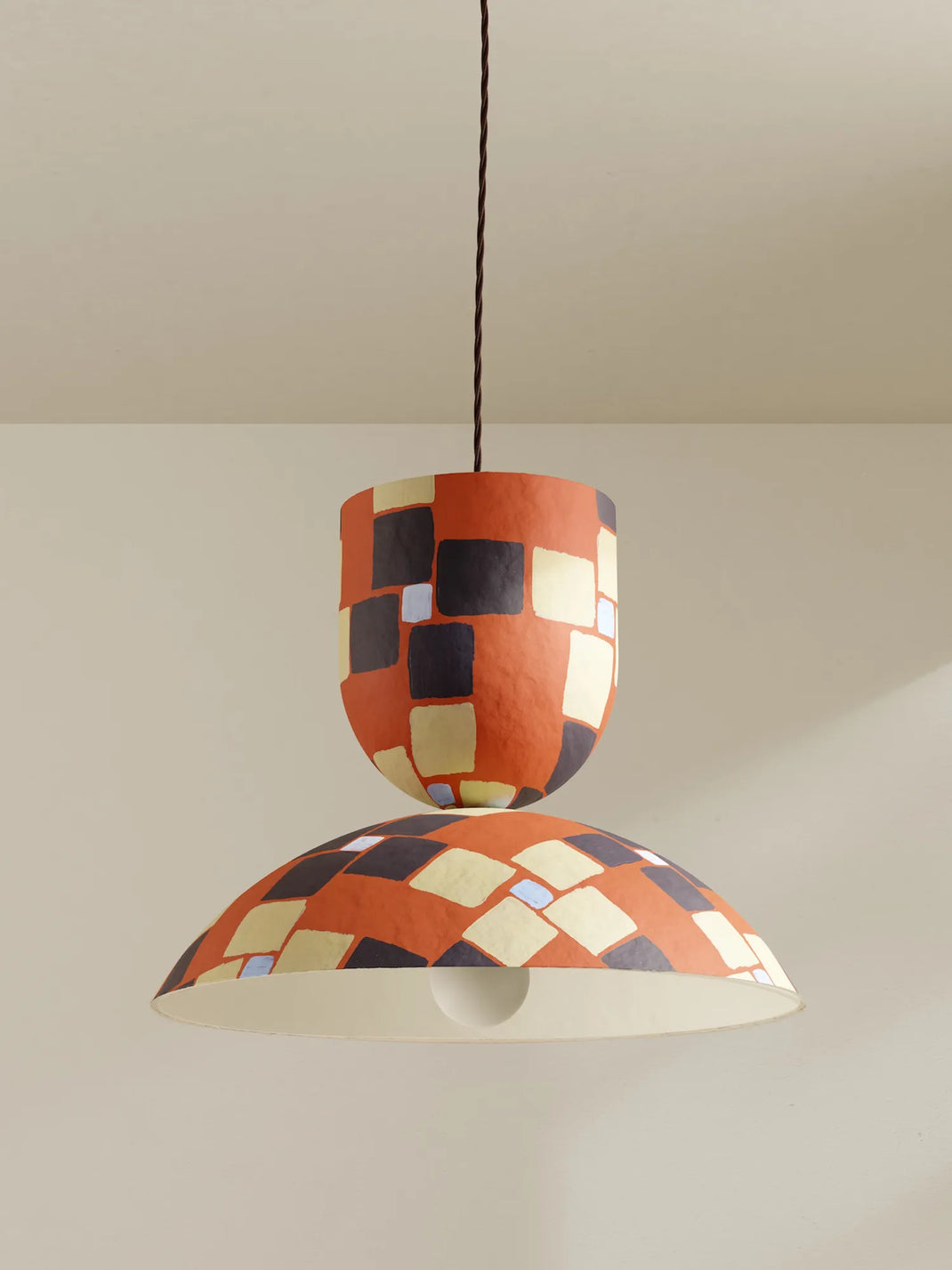 A modern two-tiered pendant light with a textured terracotta orange shade featuring a black and cream geometric checkered pattern, suspended by a brown cord against a neutral beige wall.