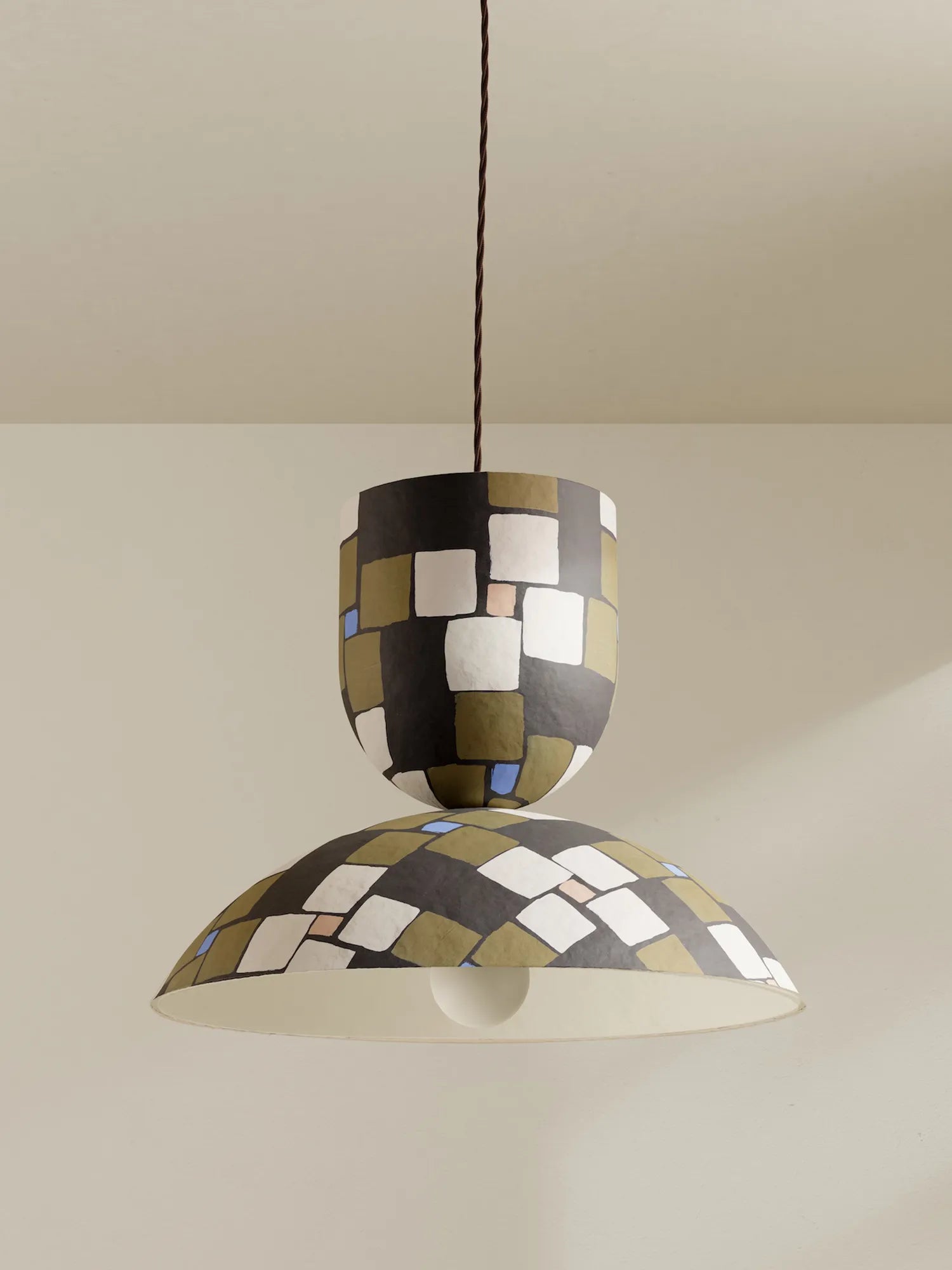 A modern pendant light with a two-tiered design, featuring a glossy black, beige, and white geometric mosaic pattern on the shades, suspended by a brown cord against a warm beige wall.