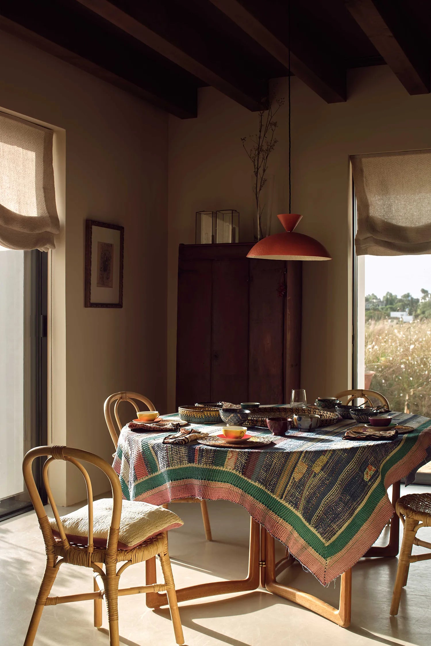A rustic dining table set with a colorful patterned tablecloth, wooden chairs, and a red pendant light in a sunlit room with dark wood ceiling beams.