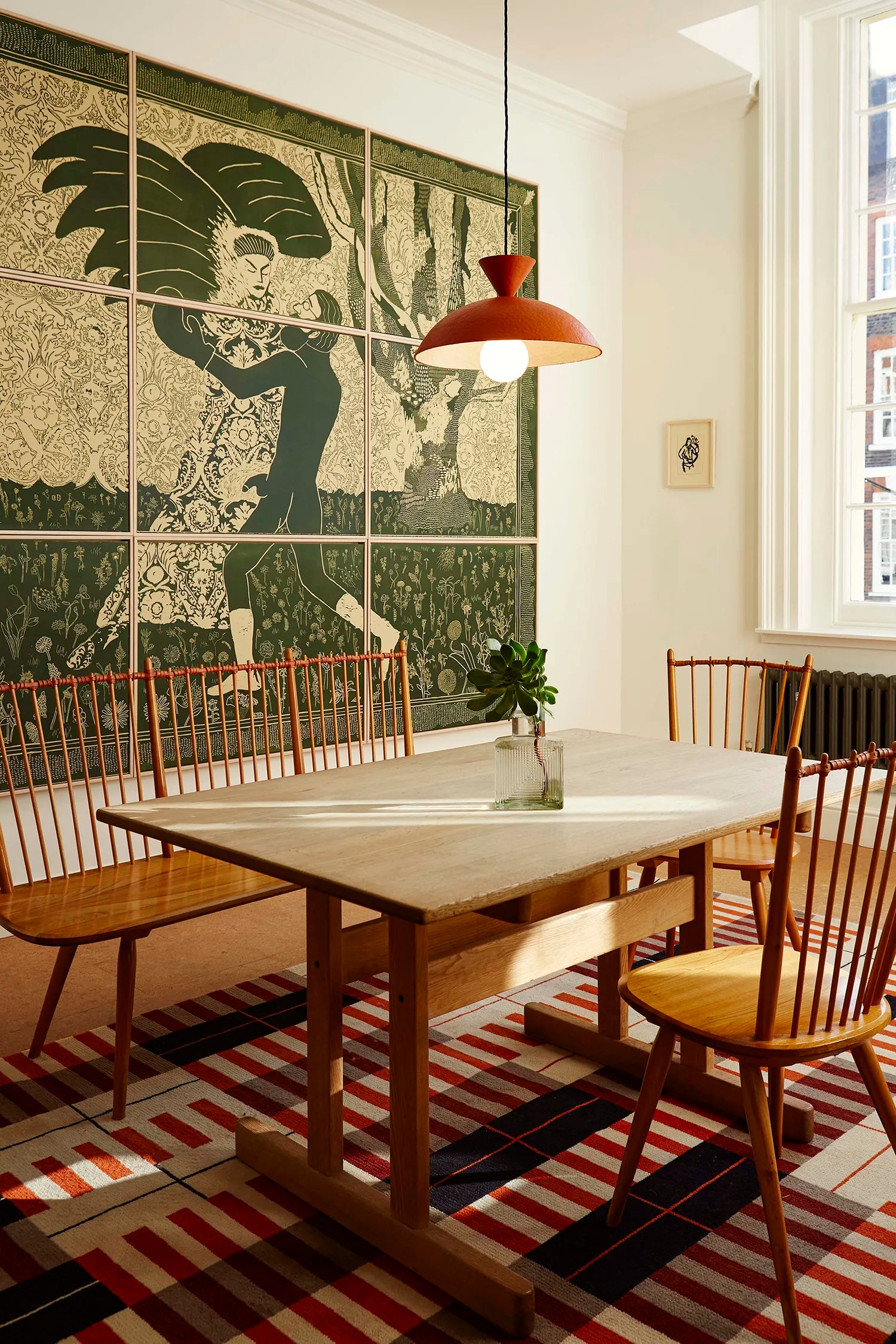 A dining room corner featuring a mid-century modern dining table and chairs, set on a striped rug. A large green and beige art deco style wall mural is visible in the background, with a terracotta-colored pendant light hanging in front.