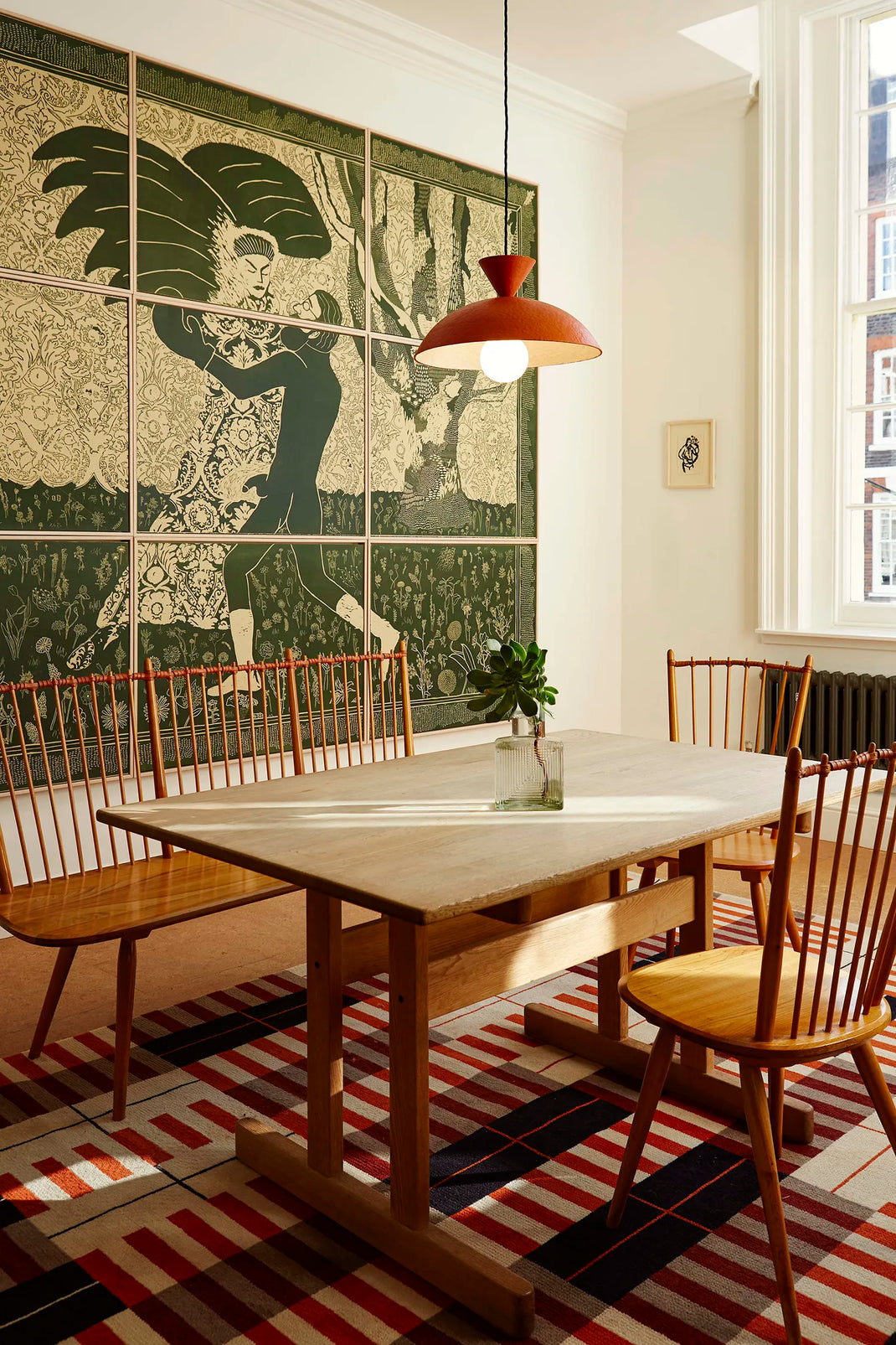 A styled dining room corner featuring a mid-century modern wooden dining table and spindle-back chairs, set on a red, black, and white striped rug. A large green and beige art deco style wall mural is visible in the background, with a terracotta-colored pendant light hanging above.