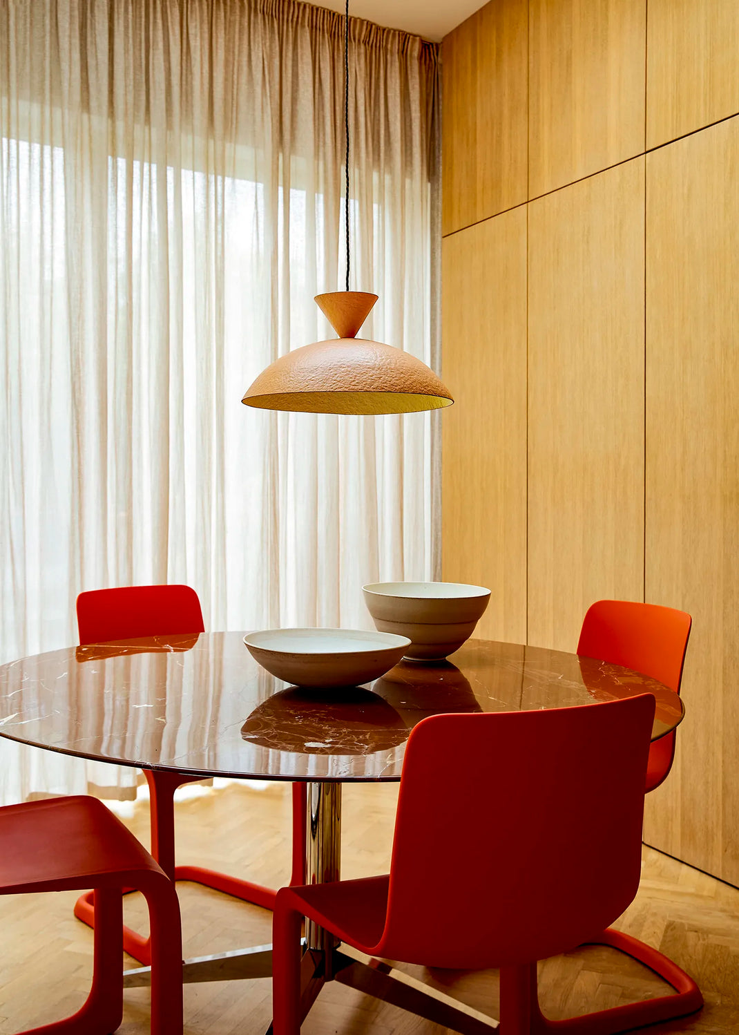 A modern dining room scene featuring a round glass-top table with four red chairs, a textured peach-coloured pendant light hanging above, and a light wood paneled wall in the background.