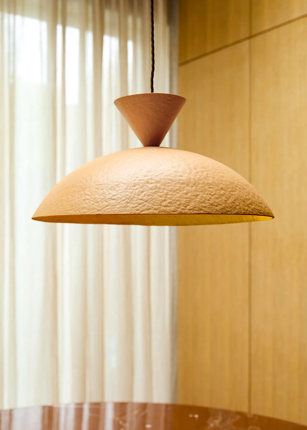 A modern pendant light with a textured, dome-shaped shade and a conical top, hanging in a warm, softly lit interior with wood paneling and sheer curtains.