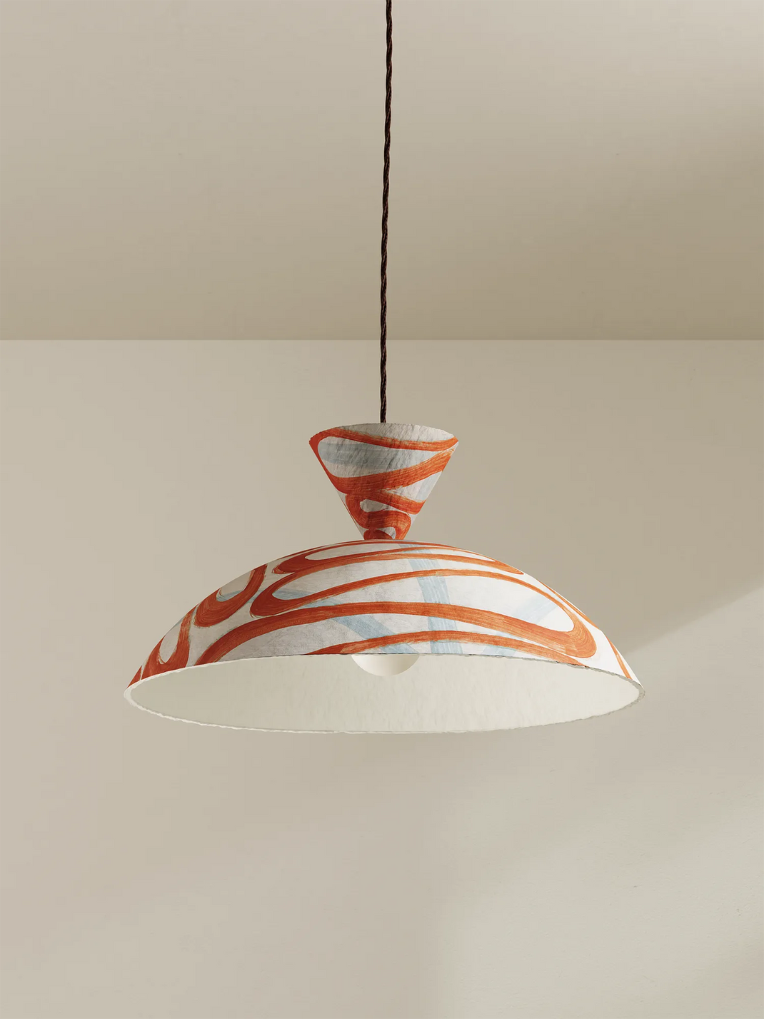 A modern pendant light with a wide, shallow dome shade featuring a white and orange marbled pattern, suspended by a brown cord against a neutral beige wall and ceiling.