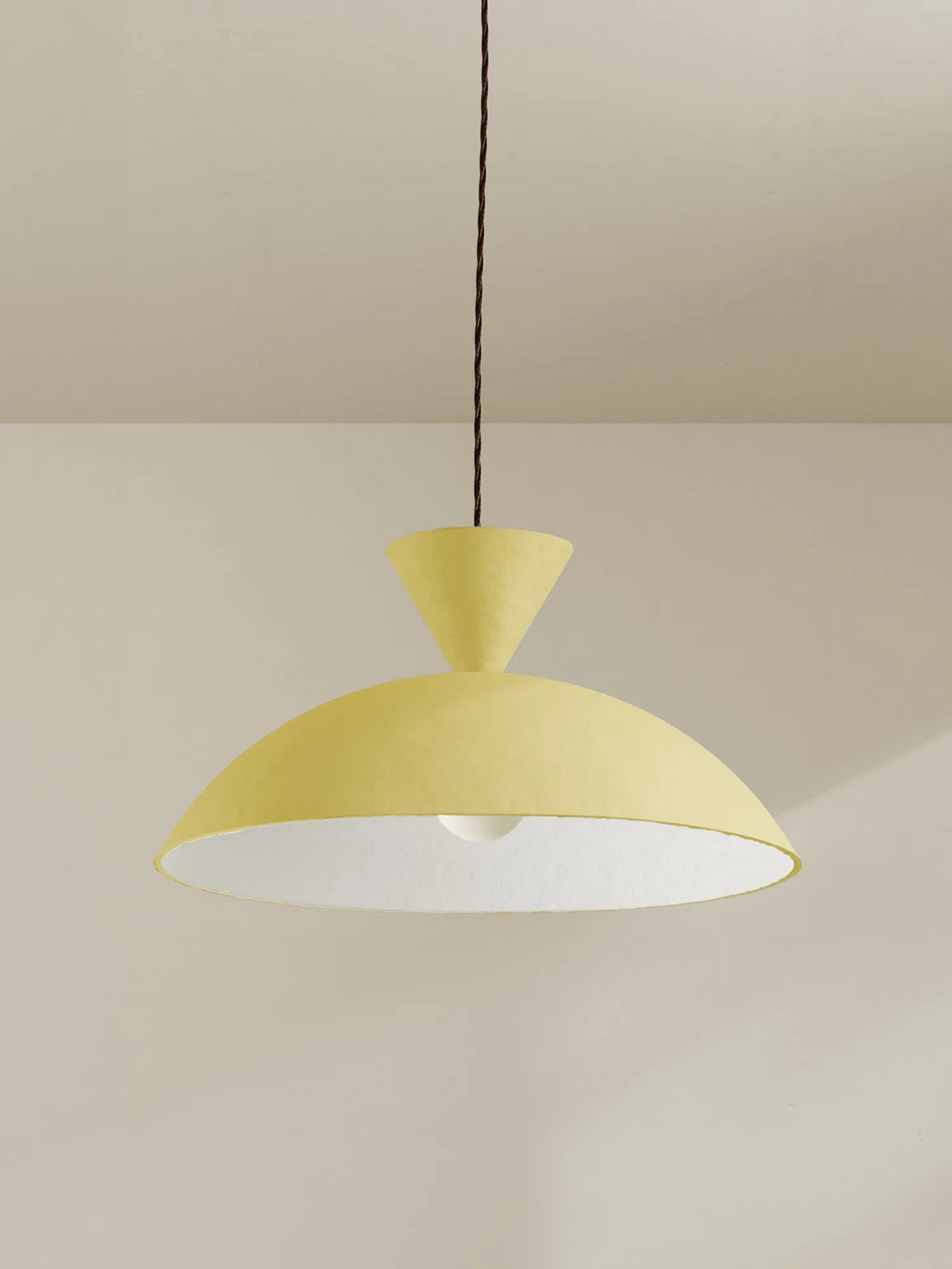 A modern pendant light with a textured pale yellow dome shade and a white interior, suspended by a brown cord against a warm beige wall and ceiling.
