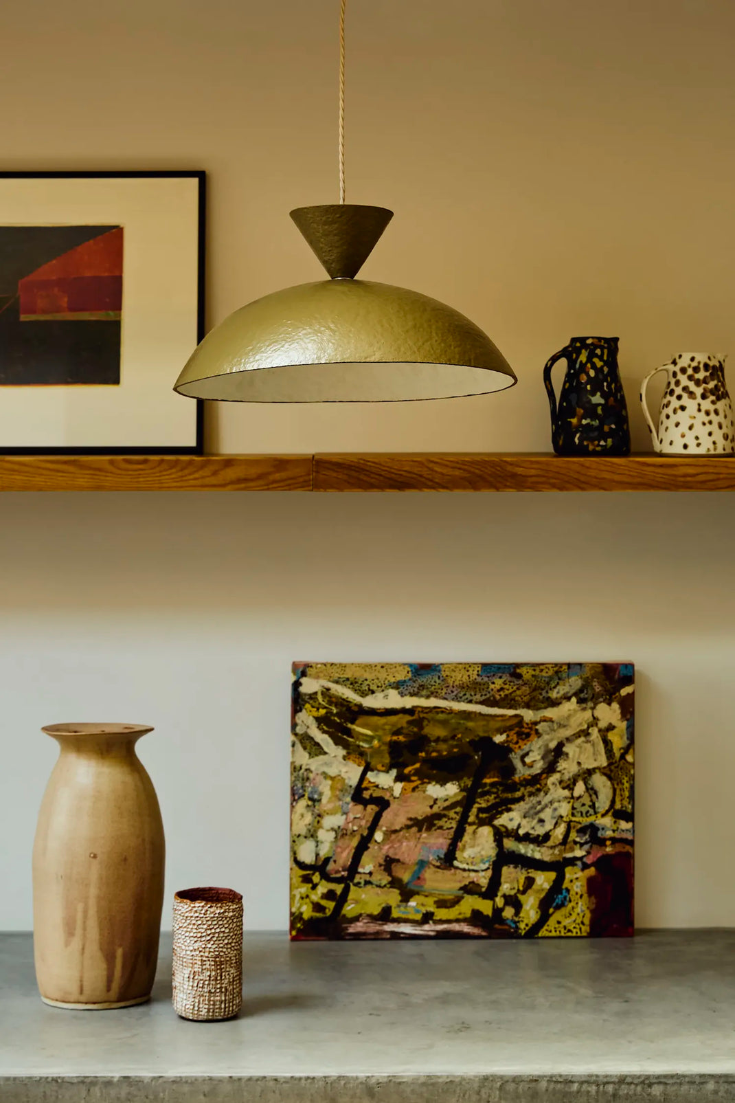 A modern pendant light with a textured green dome shade hangs on a beige wall above a wooden shelf. The shelf holds a framed abstract print, a dark patterned pitcher, and a white speckled pitcher.