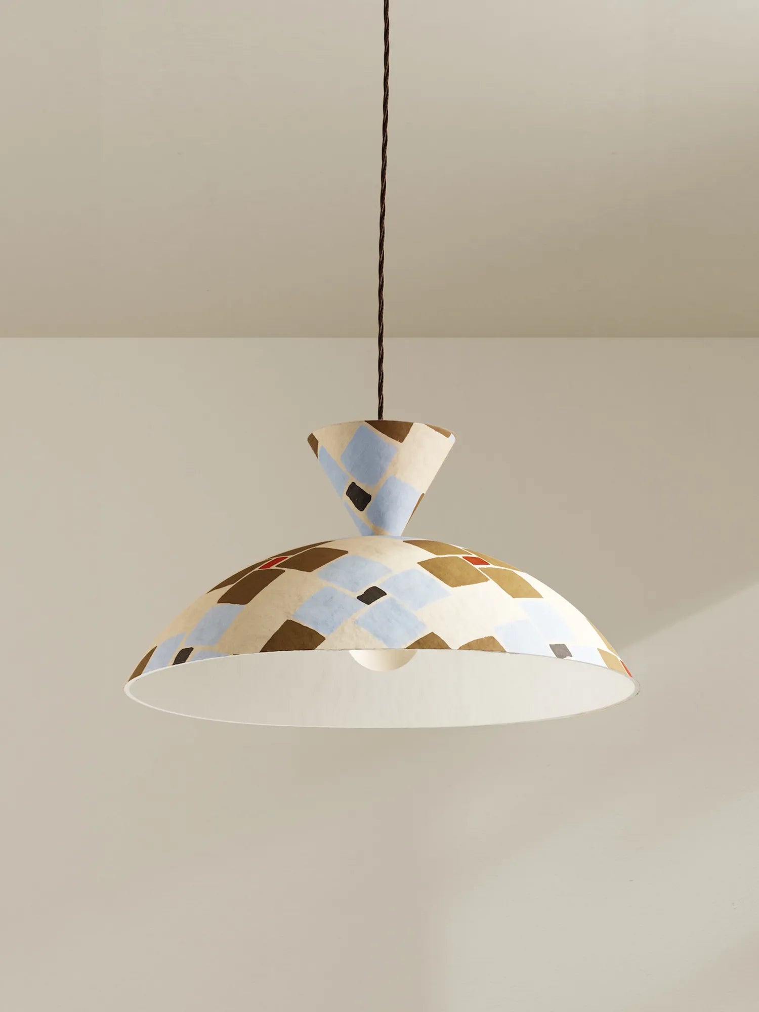 A modern pendant light with a conical shade and a wide dome shade, both decorated with a geometric pattern of beige, light blue, brown, and black shapes. The lamp is suspended by a brown cord against a neutral beige wall and ceiling.