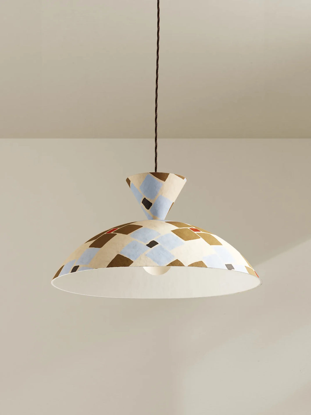 A modern pendant light with a conical shade and a wide dome shade, both decorated with a geometric pattern of beige, light blue, brown, and black shapes. The lamp is suspended by a brown cord against a neutral beige wall and ceiling.