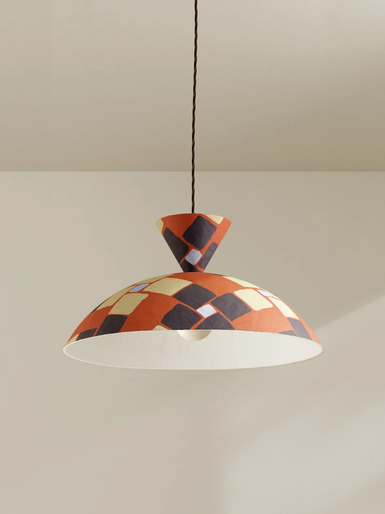 A modern pendant light with a wide dome shade featuring a geometric orange, black, and beige checkerboard pattern, suspended by a brown cord against a neutral beige wall.
