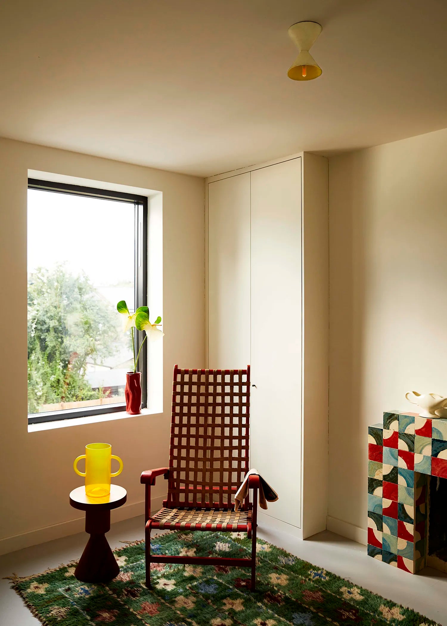 A vintage-style wooden lattice armchair with a woven seat, positioned in a room corner next to a window and a colorful geometric sideboard. A white ceiling mounted light hangs above.