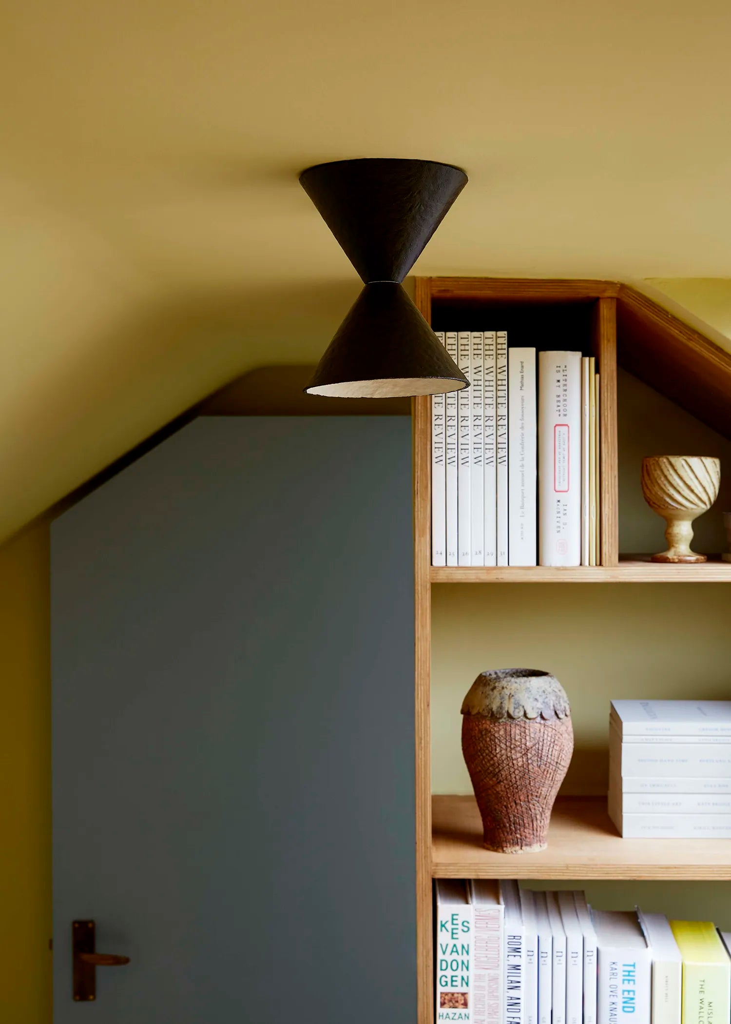 A modern brown-black cone-shaped light hanging from the ceiling in a cozy room with a slanted roof, next to a wooden bookshelf filled with books and decorative vases.