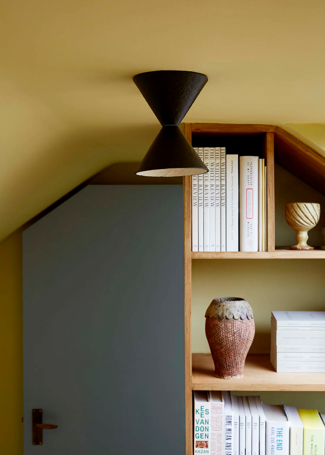 A modern brown-black cone-shaped light hanging from the ceiling in a cozy room with a slanted roof, next to a wooden bookshelf filled with books and decorative vases.