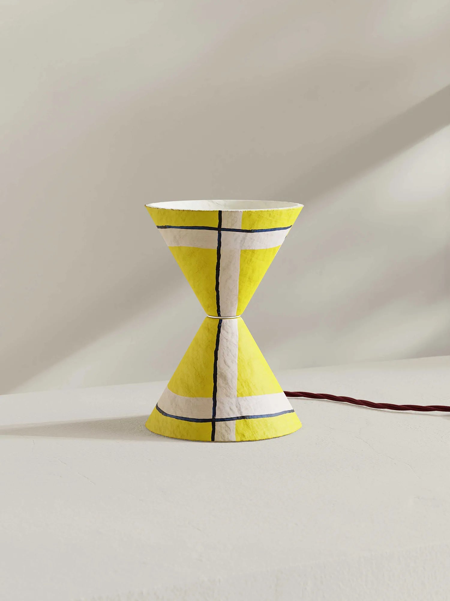 A modern hourglass-shaped table lamp with a textured yellow, white, and navy blue pattern, shown on a minimalist beige background with a dark red power cord.