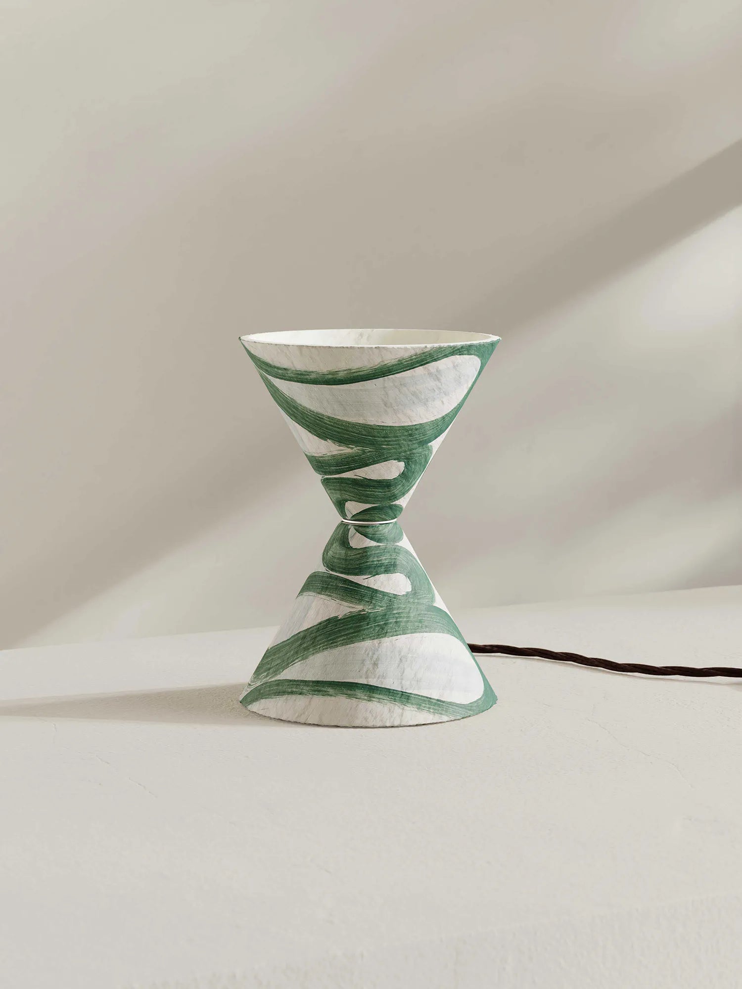 A sculptural hourglass-shaped table lamp with a green and white pattern, set against a minimalist beige background with a soft shadow.