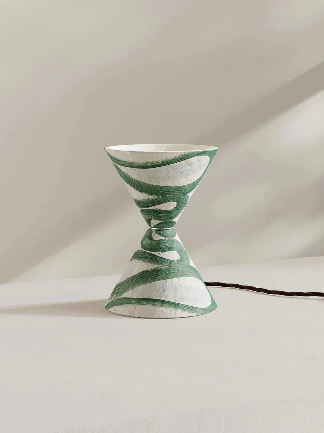 A sculptural hourglass-shaped table lamp with a green and white pattern, set against a minimalist beige background with a soft shadow.