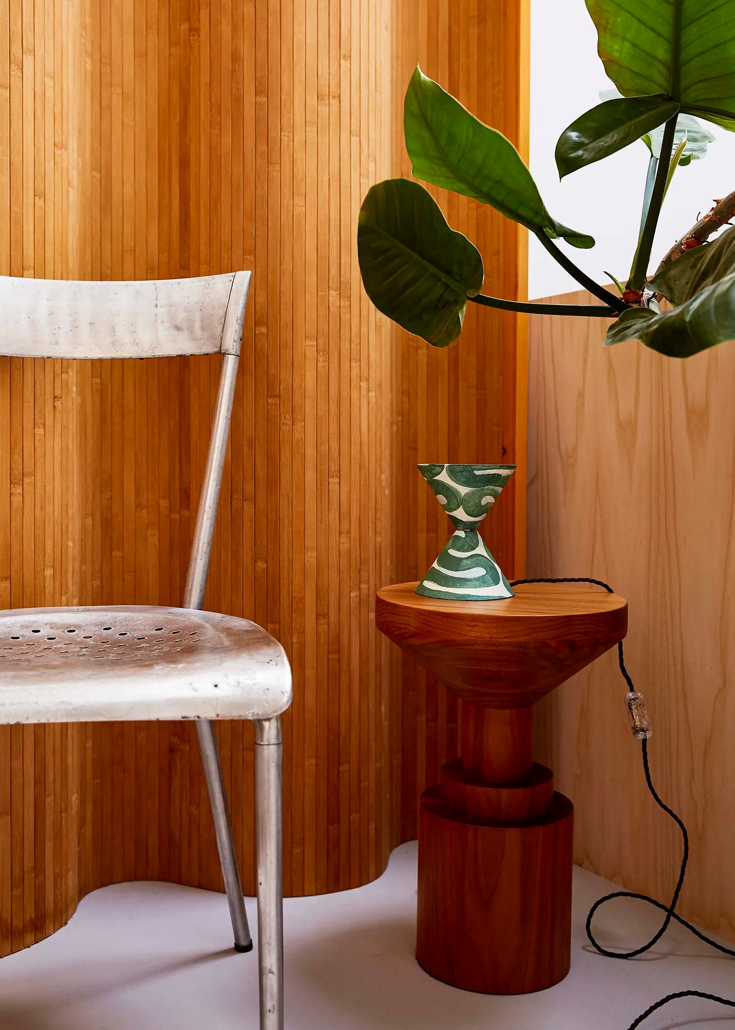 A styled interior corner featuring a metal chair on the left, a small round wooden side table holding a green and white table lamp, and a large green houseplant in the upper right against a warm wooden screen.