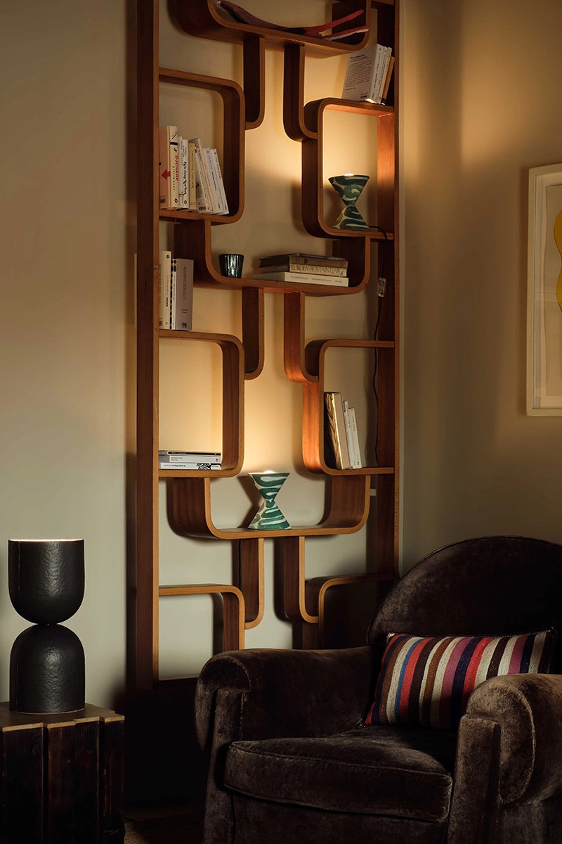 In a sitting room, two small lamps with a green pattern is on a shelf, illuminating the books above.