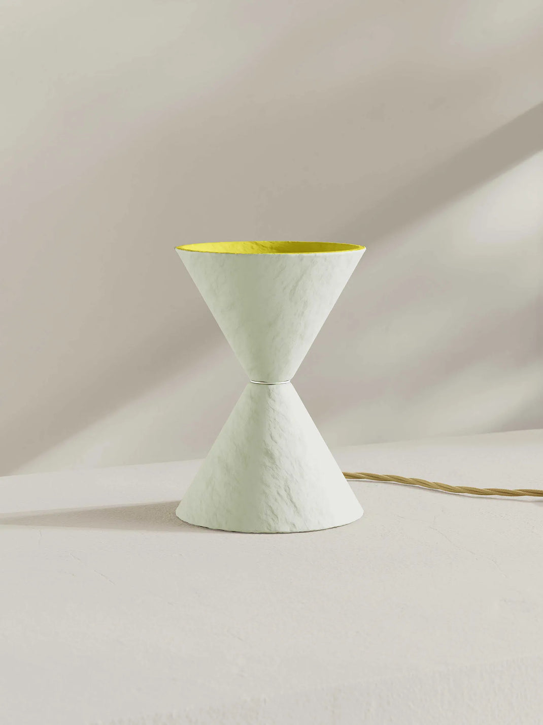 A sculptural hourglass-shaped table lamp with a textured off-white exterior and a bright yellow interior, shown on a neutral cream surface with a gold-toned power cord.