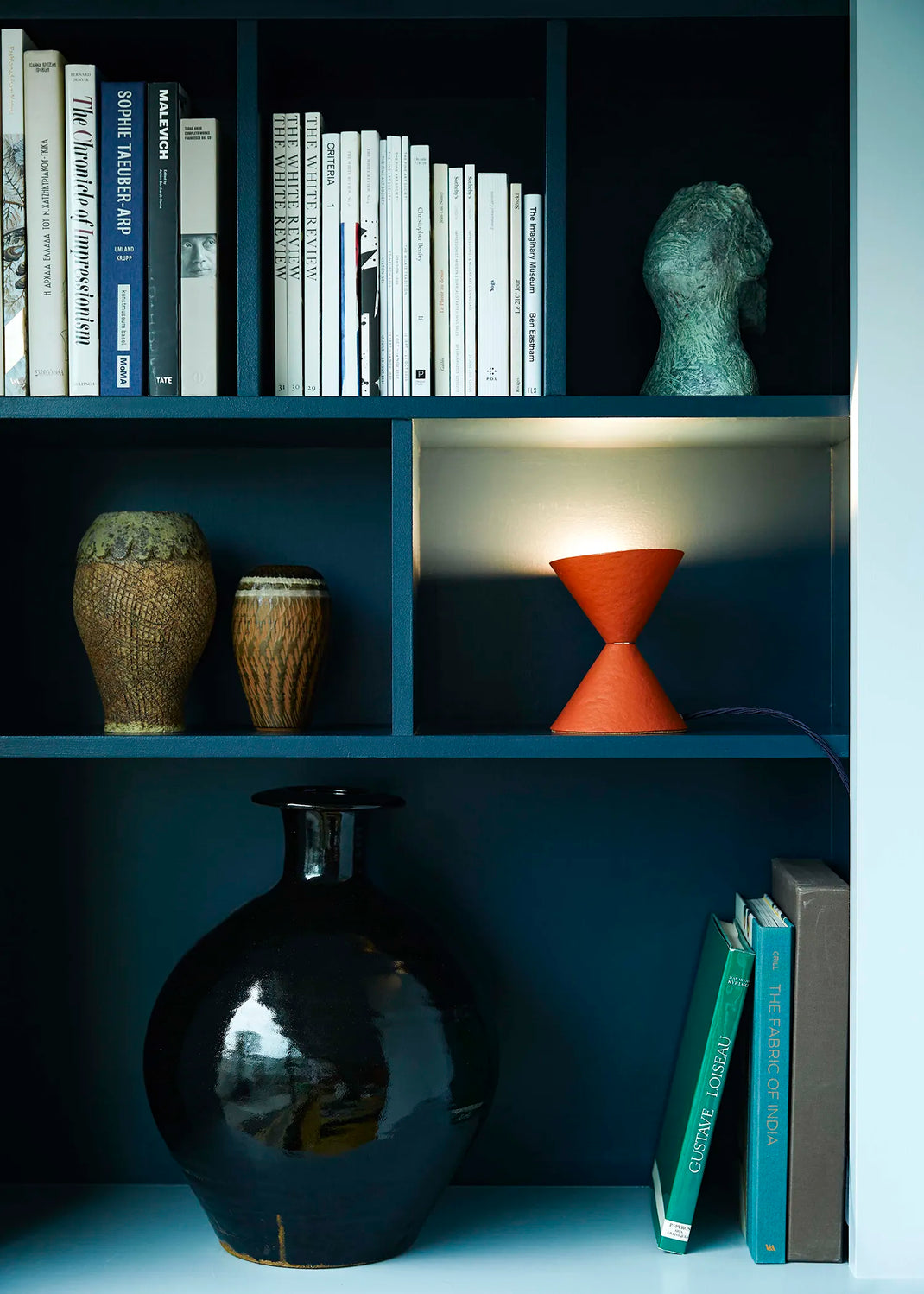 A styled interior shelf scene with dark blue shelving holding books, vases, a sculpture, and a terracotta-colored hourglass-shaped lamp illuminated from within.