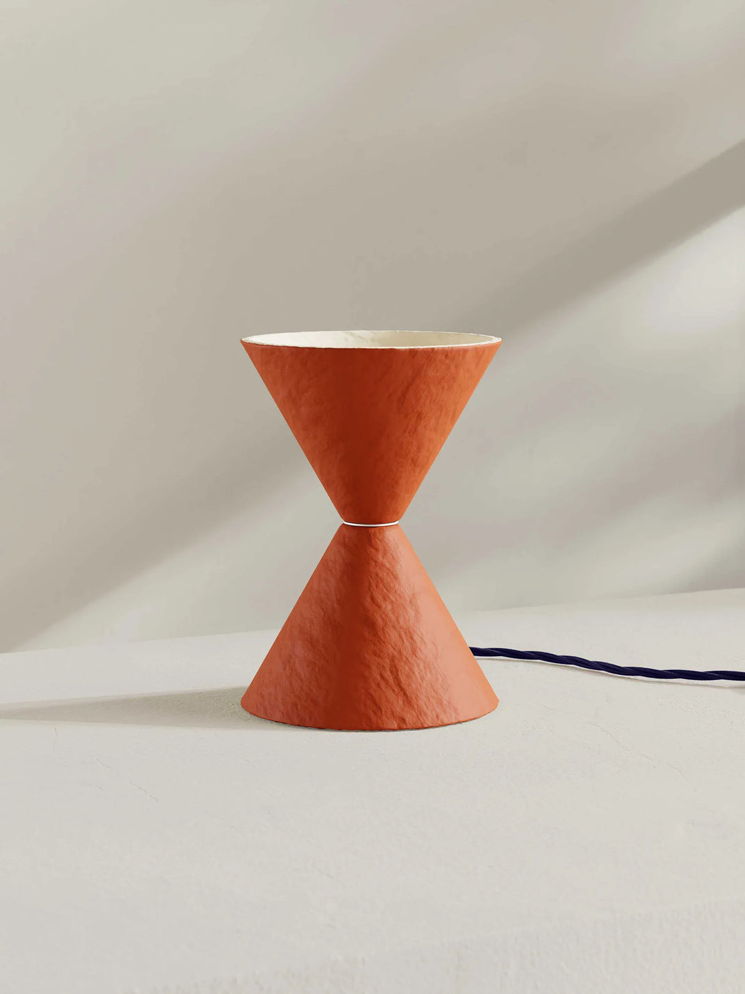 A sculptural table lamp with a textured terracotta orange finish, featuring a double-cone hourglass design with a white interior, set against a minimalist off-white background.