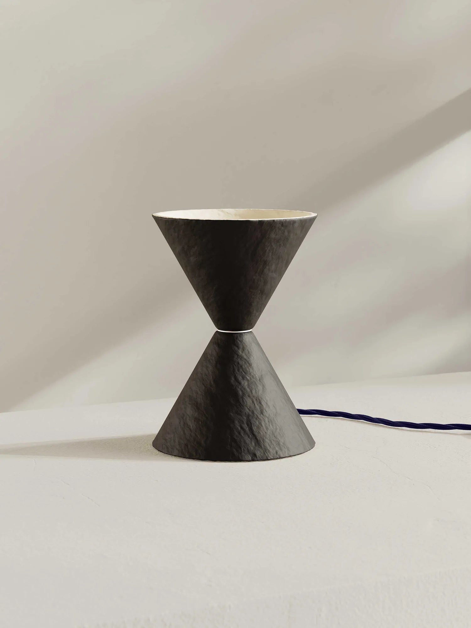A sculptural hourglass-shaped table lamp with a brown-black textured exterior and a light cream interior, shown on a minimalist off-white surface with a dark blue cord running to the right.