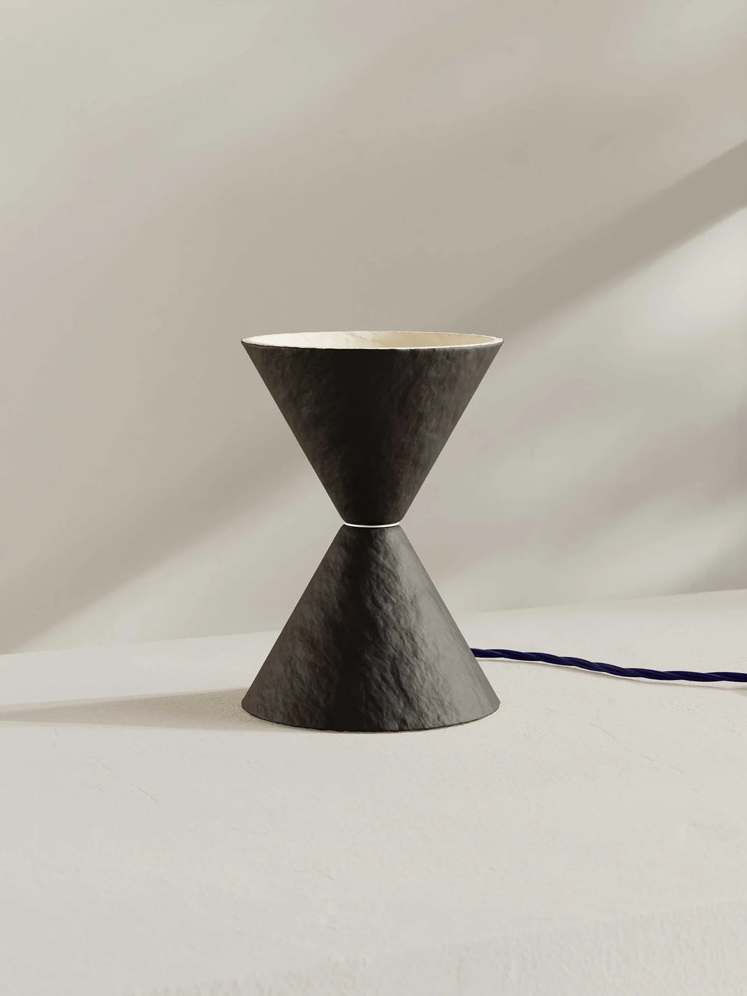 A sculptural hourglass-shaped table lamp with a brown-black textured exterior and a light cream interior, shown on a minimalist off-white surface with a dark blue cord running to the right.
