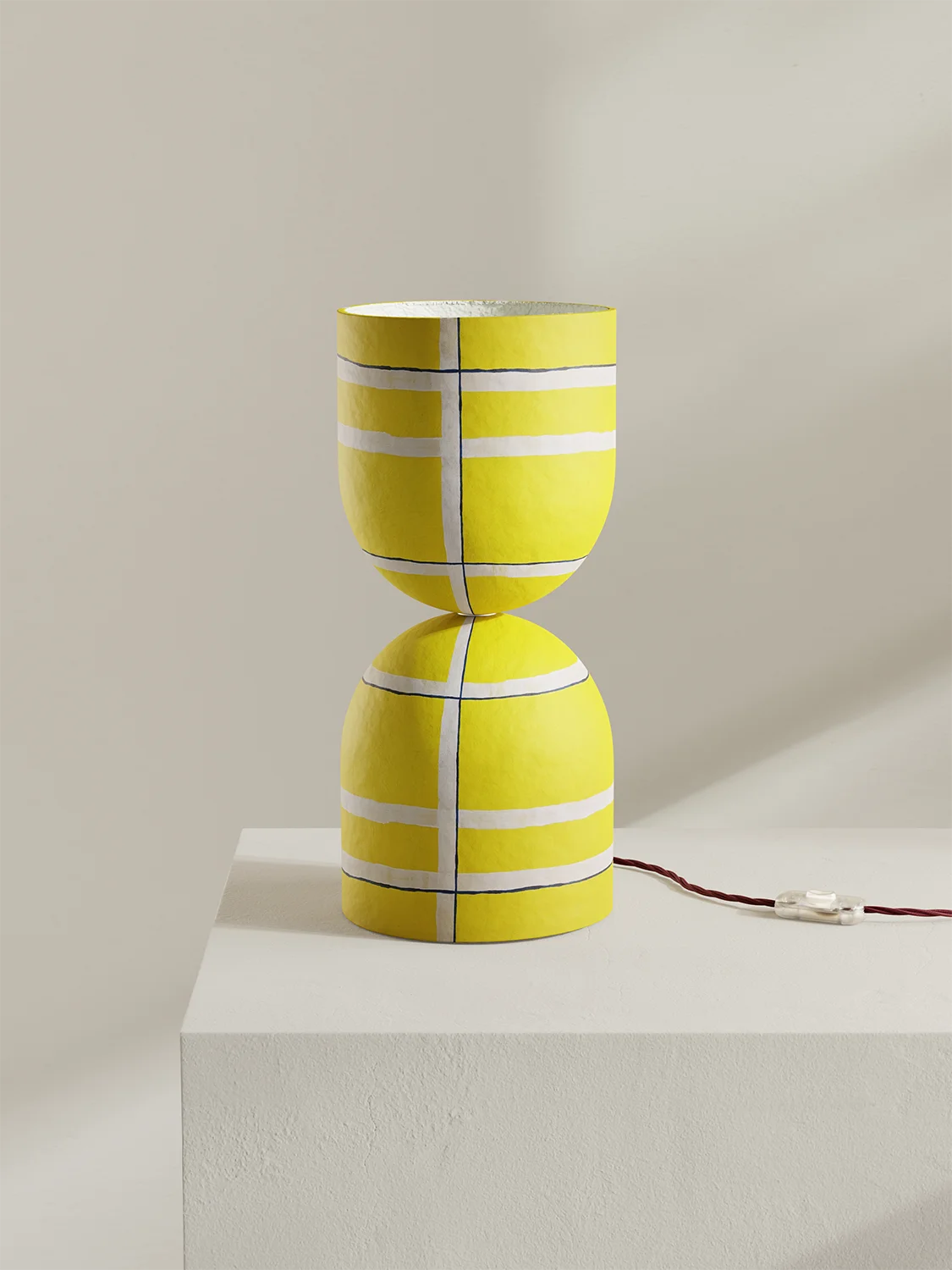 A bright yellow table lamp with a sculptural hourglass-like form and white painted stripe accents, displayed on a white pedestal against a warm beige wall. A dark red power cord extends from the base.