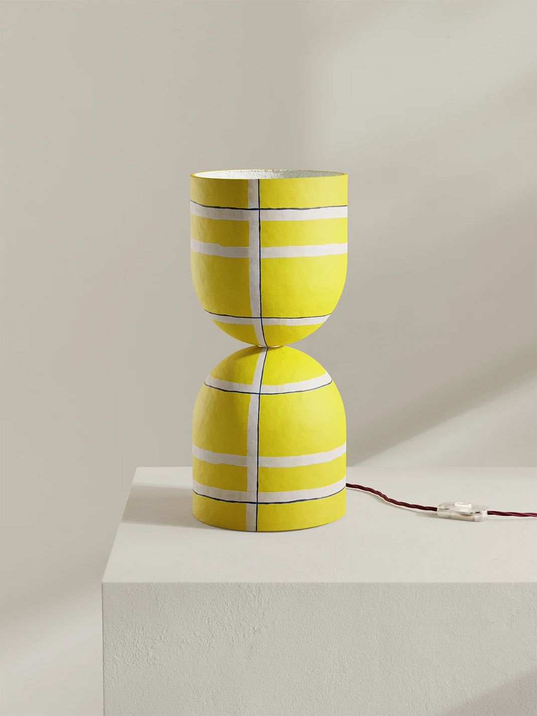 A bright yellow table lamp with a sculptural hourglass-like form and white painted stripe accents, displayed on a white pedestal against a warm beige wall. A dark red power cord extends from the base.