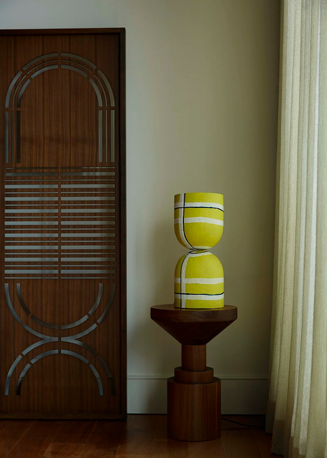 A yellow and white striped hourglass-shaped table lamp sits on a dark wooden pedestal side table in a room corner next to a cream curtain.