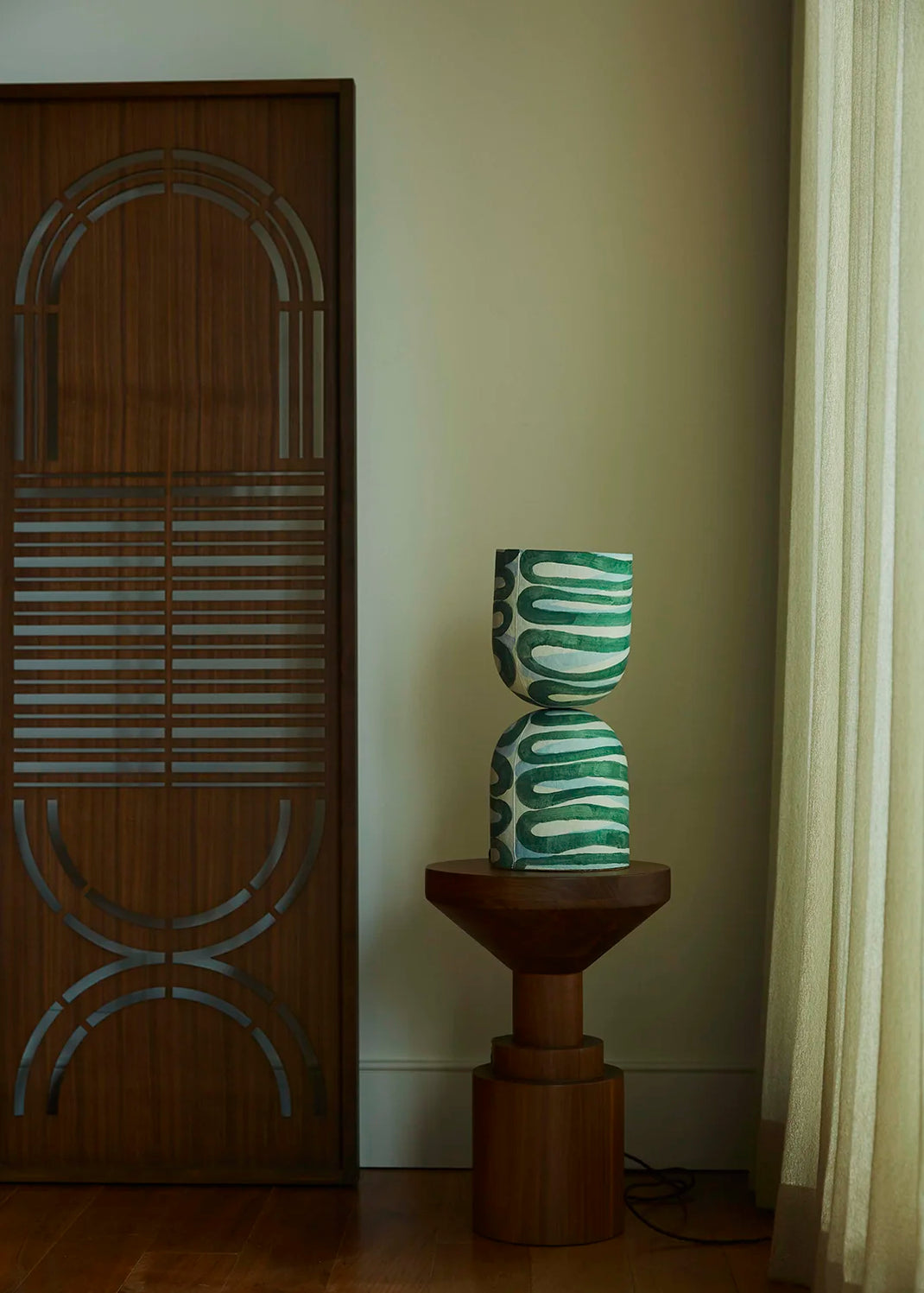 A sculptural hourglass-shaped table lamp with a green and white wavy pattern sits on a wooden pedestal side table in a room corner next to a wooden door and curtain.