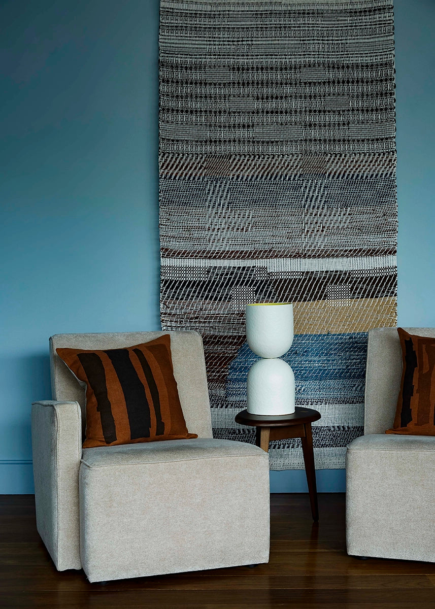 A white table lamp in the middle of two beige armchairs with brown striped cushions in a room with blue walls and a textured wall hanging.