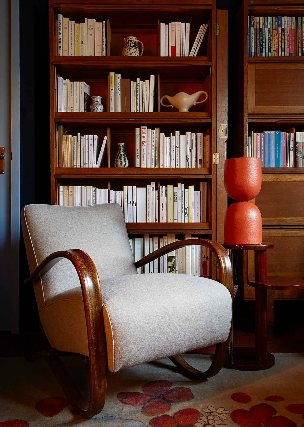 White armchair in a room with wooden bookshelves filled with books and decorative items. An orange-red table lamp sits on a wooden table beside the armchair.