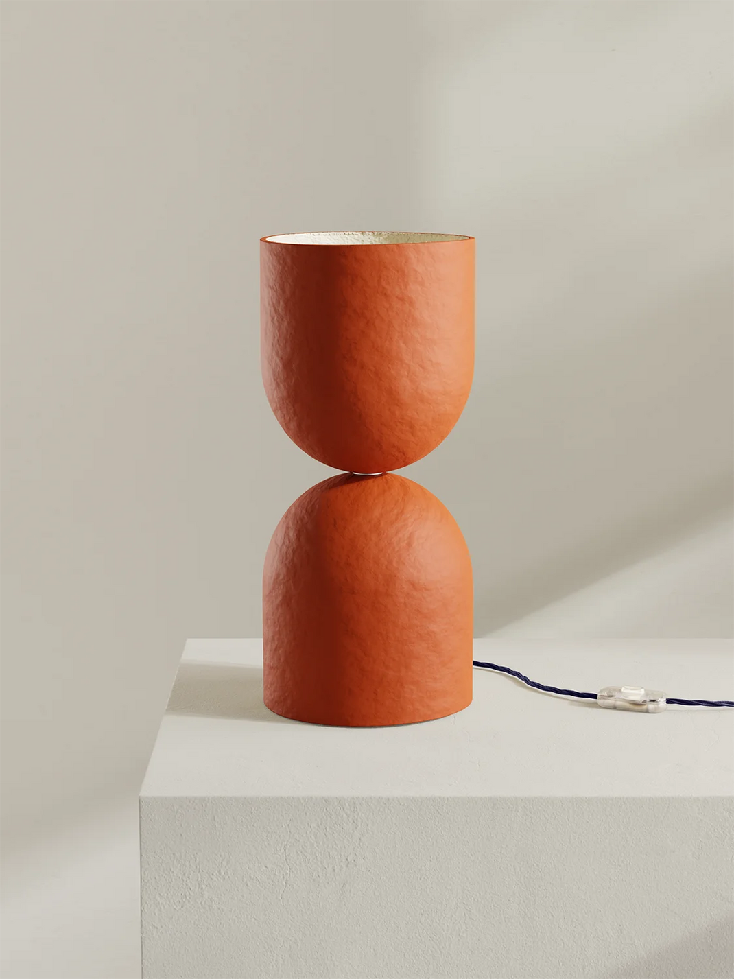 An hourglass-shaped table lamp with a textured terracotta orange finish, displayed on a white pedestal against a neutral beige background.