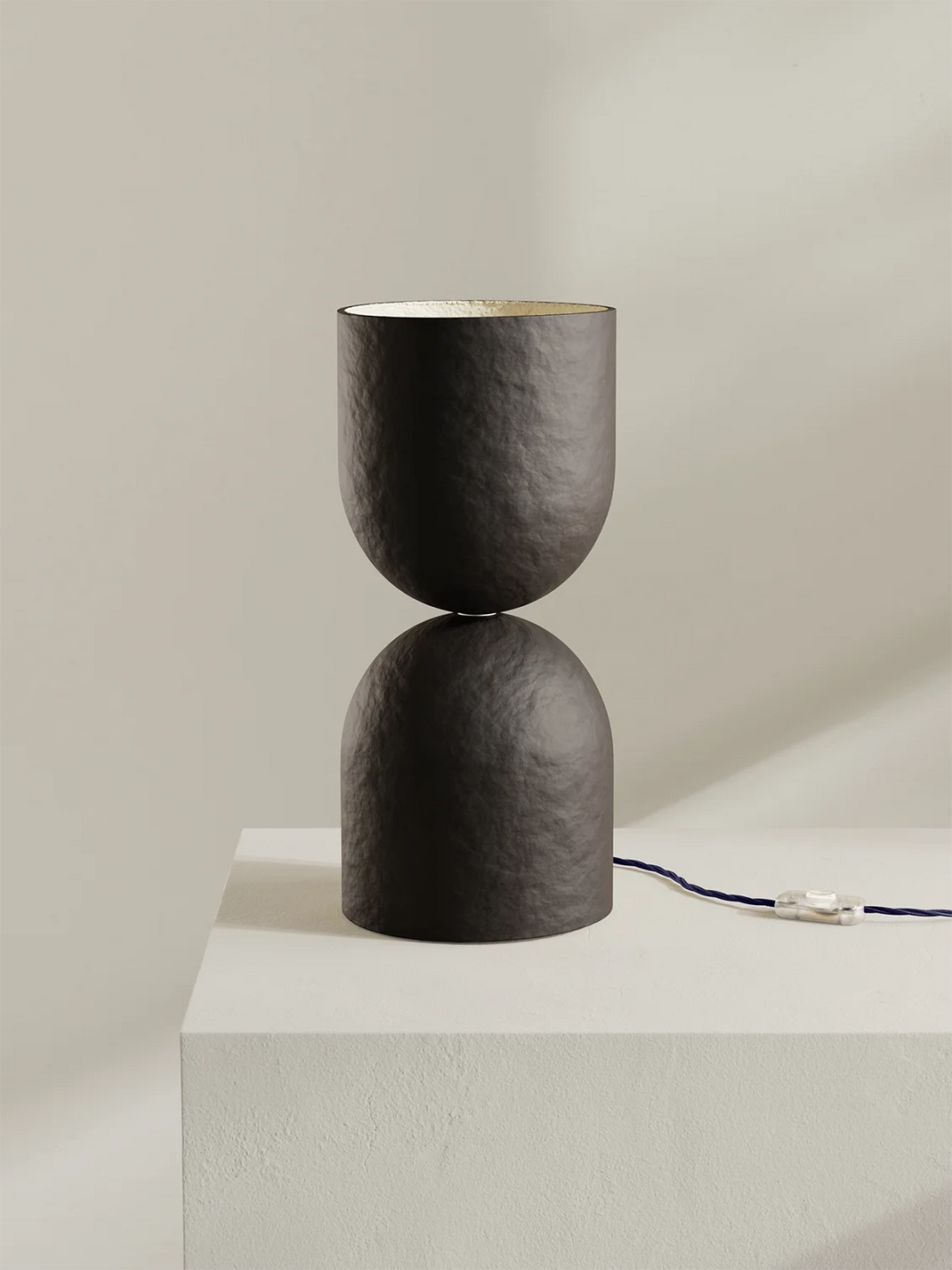 A modern table lamp with a brown-black textured finish, designed in a double-hemispherical hourglass shape, resting on a white pedestal against a warm beige background.