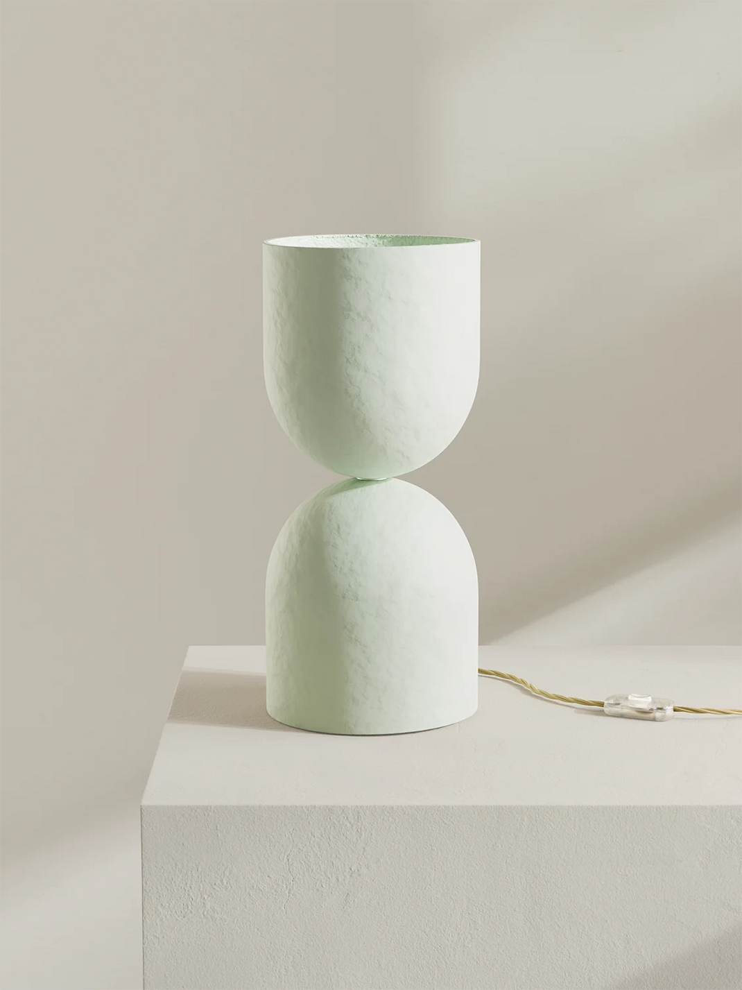 An off-white textured table lamp with an hourglass design, displayed on a white pedestal against a warm beige background.