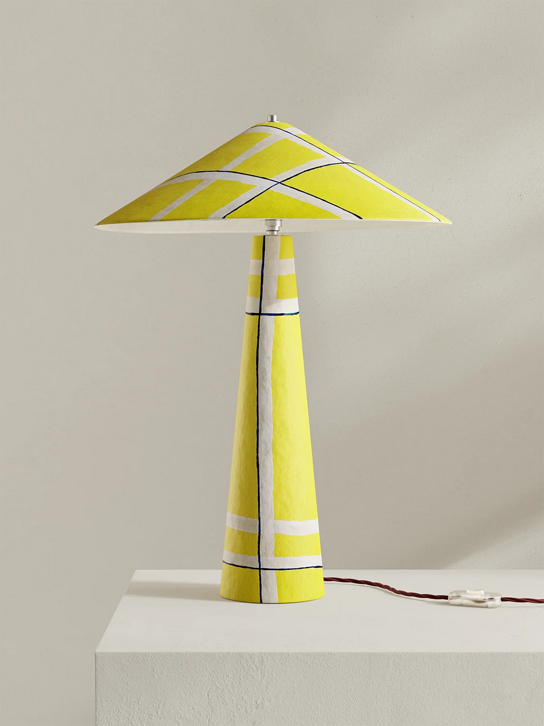 A modern table lamp with a tapered yellow ceramic base and a matching cone-shaped shade, decorated with white and navy geometric lines, set on a white pedestal against a neutral beige background.