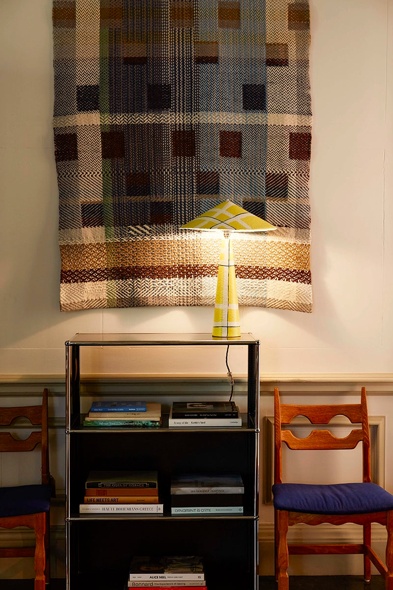 Yellow lamp on a shelf with books, against a wall with a woven tapestry.