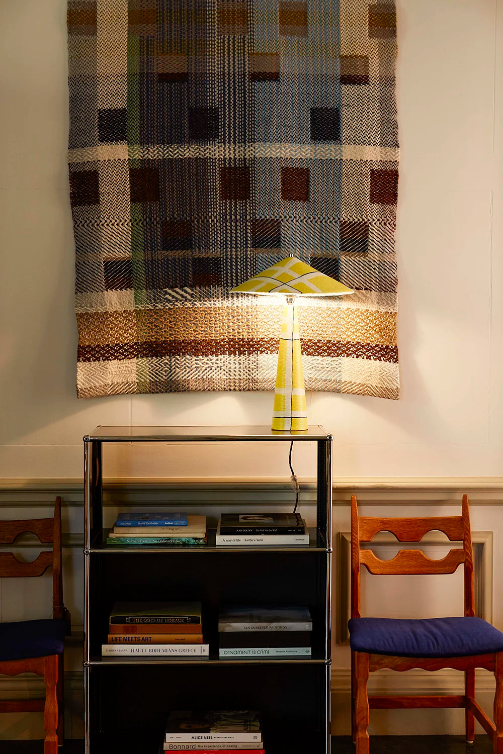 Yellow lamp on a shelf with books, against a wall with a woven tapestry.