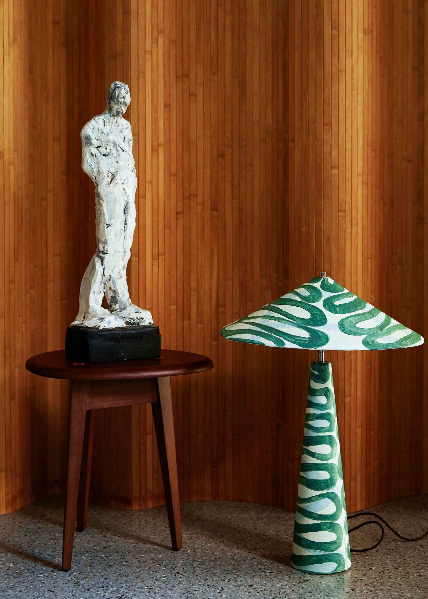 Parasol table lamp made of paper pulp in swirling green Serpent pattern, placed on the floor in front of wooden panelled screen.