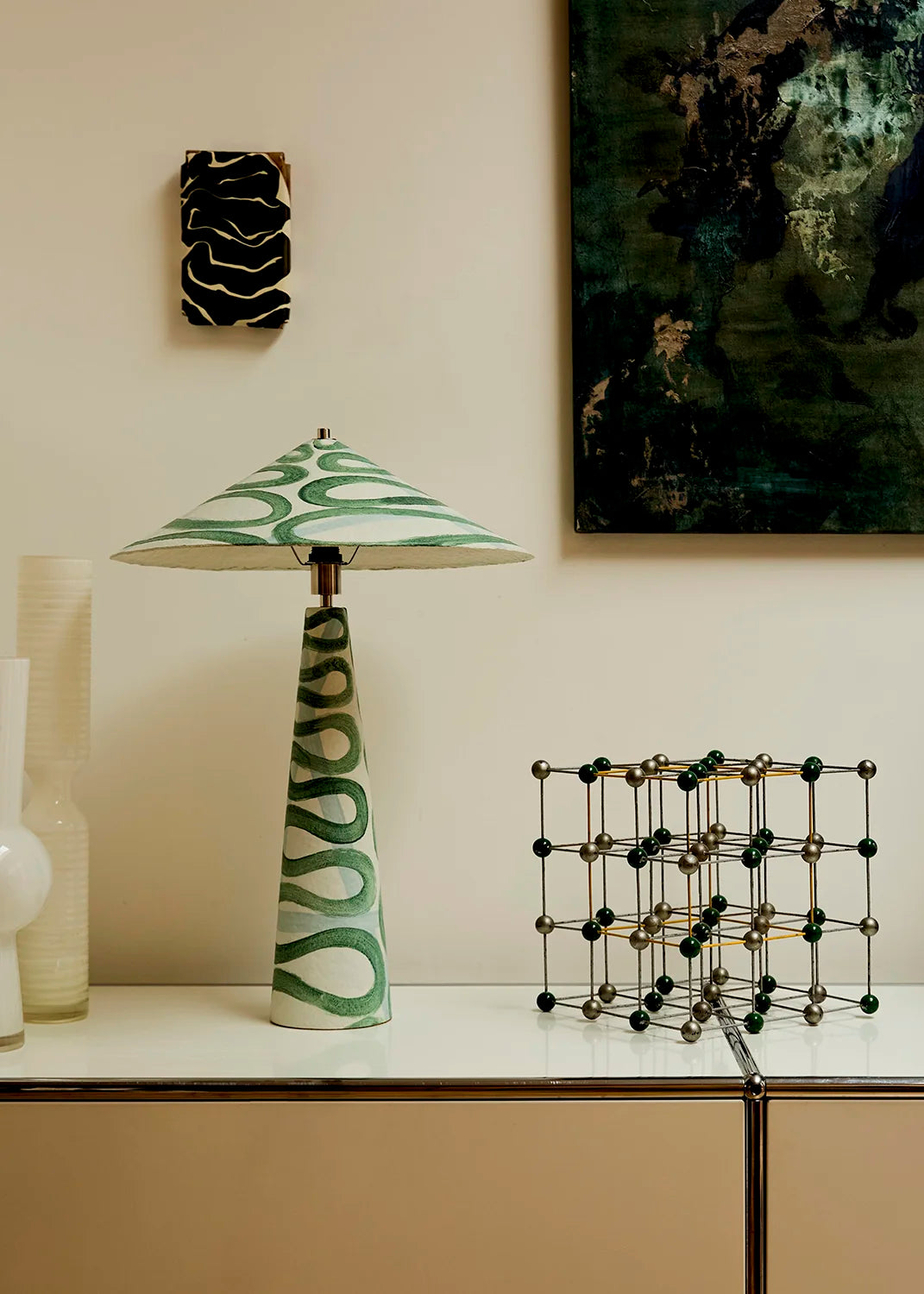A green and white patterned table lamp with a conical shade sits on a white console table against a beige wall, styled with a geometric wire sculpture and framed wall art.