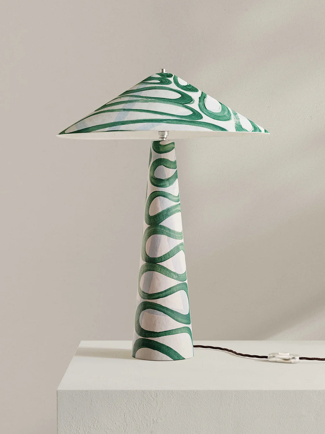 A table lamp with a tapered white base and a wide conical shade, both decorated with a green wavy, organic pattern, shown on a white pedestal against a neutral beige background.
