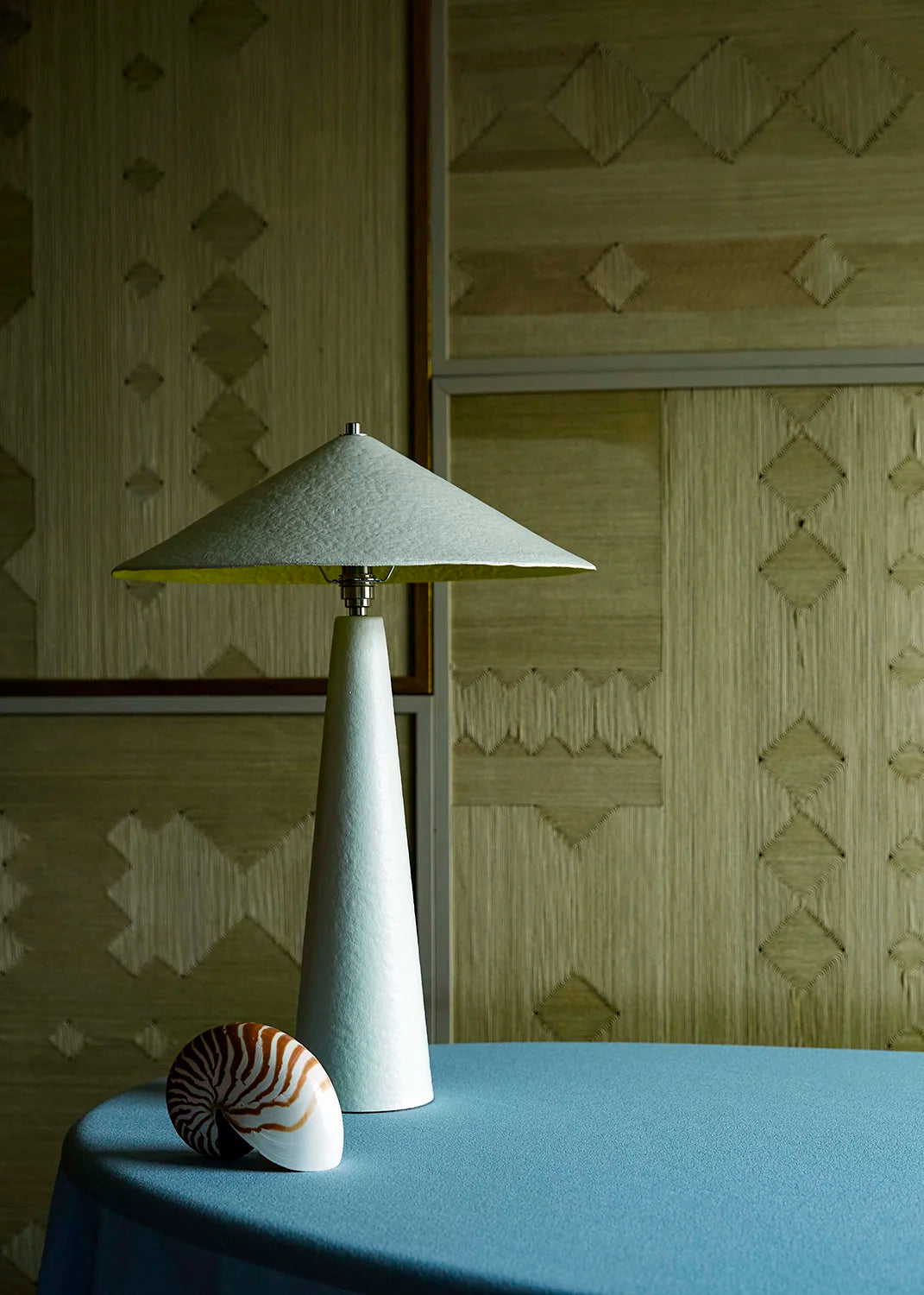 A modern table lamp with a tapered white base with a yellow interior shade sits on a blue table against a patterned beige wall.
