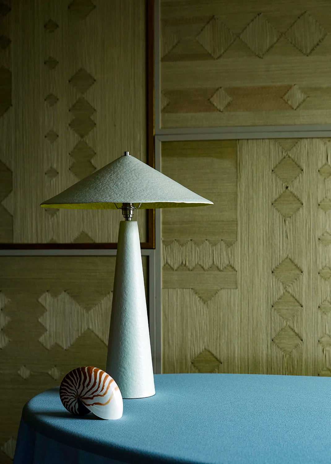 A modern table lamp with a tapered white base with a yellow interior shade sits on a blue table against a patterned beige wall.