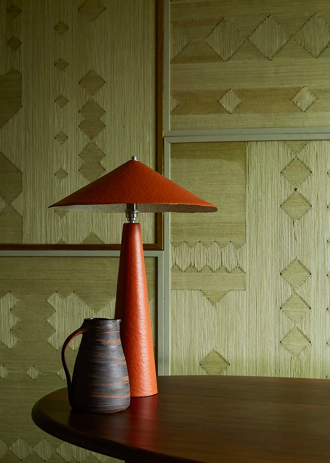 A mid-century modern table lamp with a terracotta orange conical shade and matching textured orange base, set on a dark wood table against a patterned beige wallpaper background.