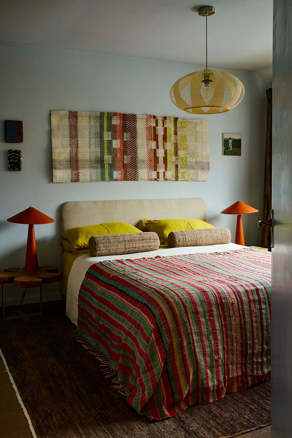 A styled bedroom interior featuring a bed with a red, green, and beige striped quilt, yellow pillows, a beige upholstered headboard, orange table lamps, and a large multicolor geometric wall hanging above.