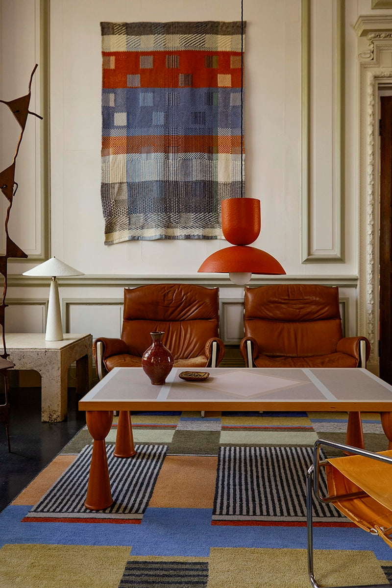 Modern living room with a textured wall hanging, white table lamp, brick red pendant, leather chairs, and a wooden table.