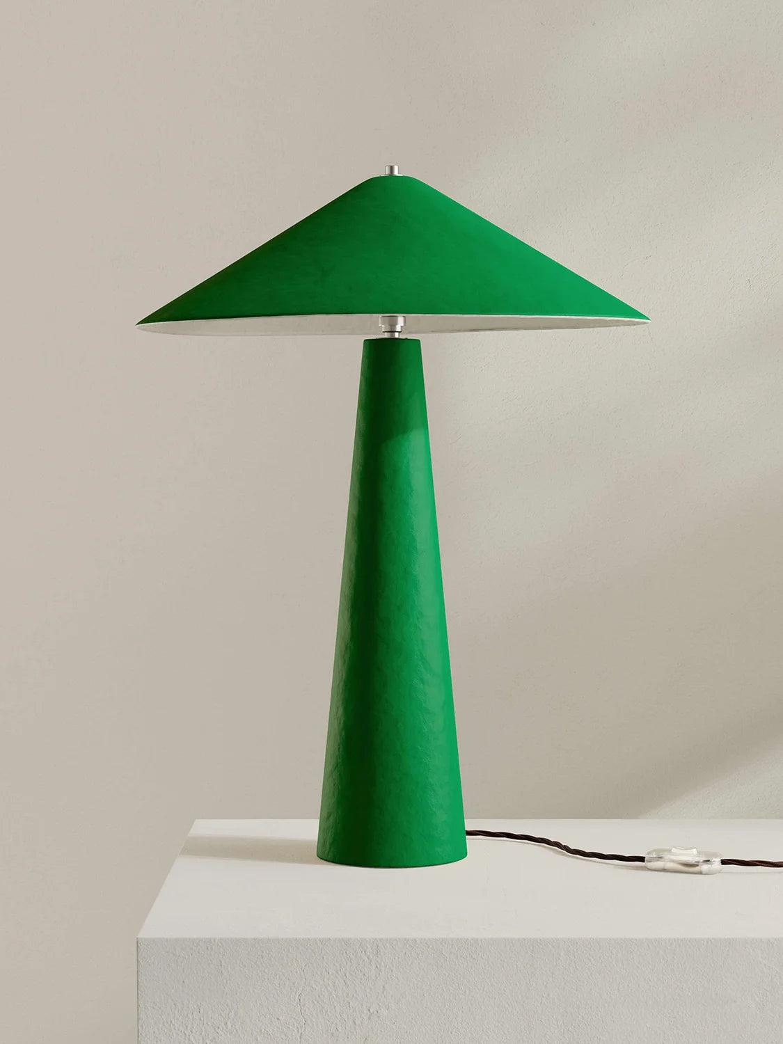 A modern green table lamp with a tapered conical shade and matching base, shown on a white pedestal against a light beige wall.