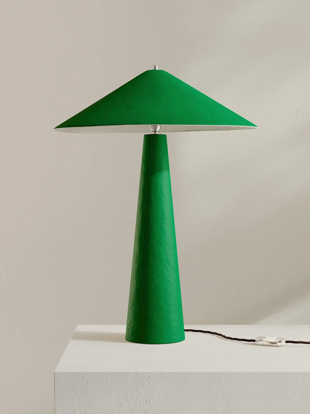 A modern green table lamp with a tapered conical shade and matching base, shown on a white pedestal against a light beige wall.