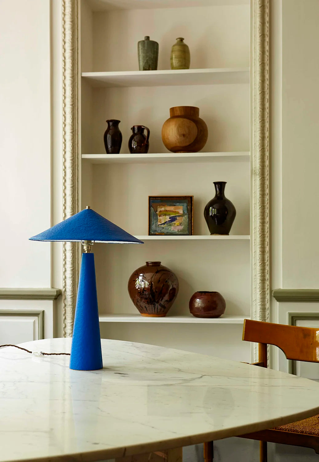 Blue table lamp on a marble table with decorative vases on shelves in the background.