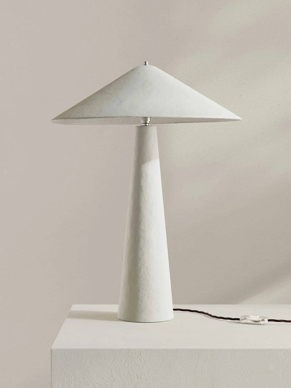 A modern table lamp with a wide, shallow conical shade and a tapered cylindrical base, both in a textured white finish, displayed on a matching pedestal against a neutral beige background.